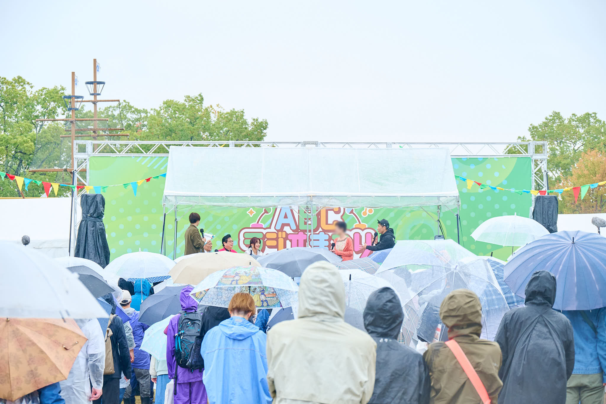ABCラジオまつりのステージ前で傘を差した観客が集まる様子、緑の背景にイベントロゴ