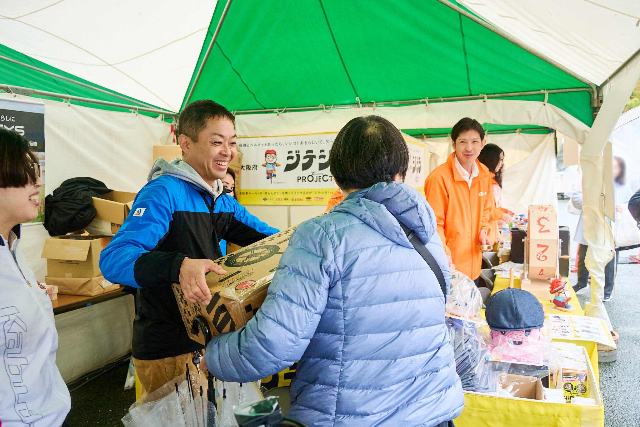 イベント会場のテントで景品を受け取る参加者とスタッフ、段ボール箱が並ぶ様子