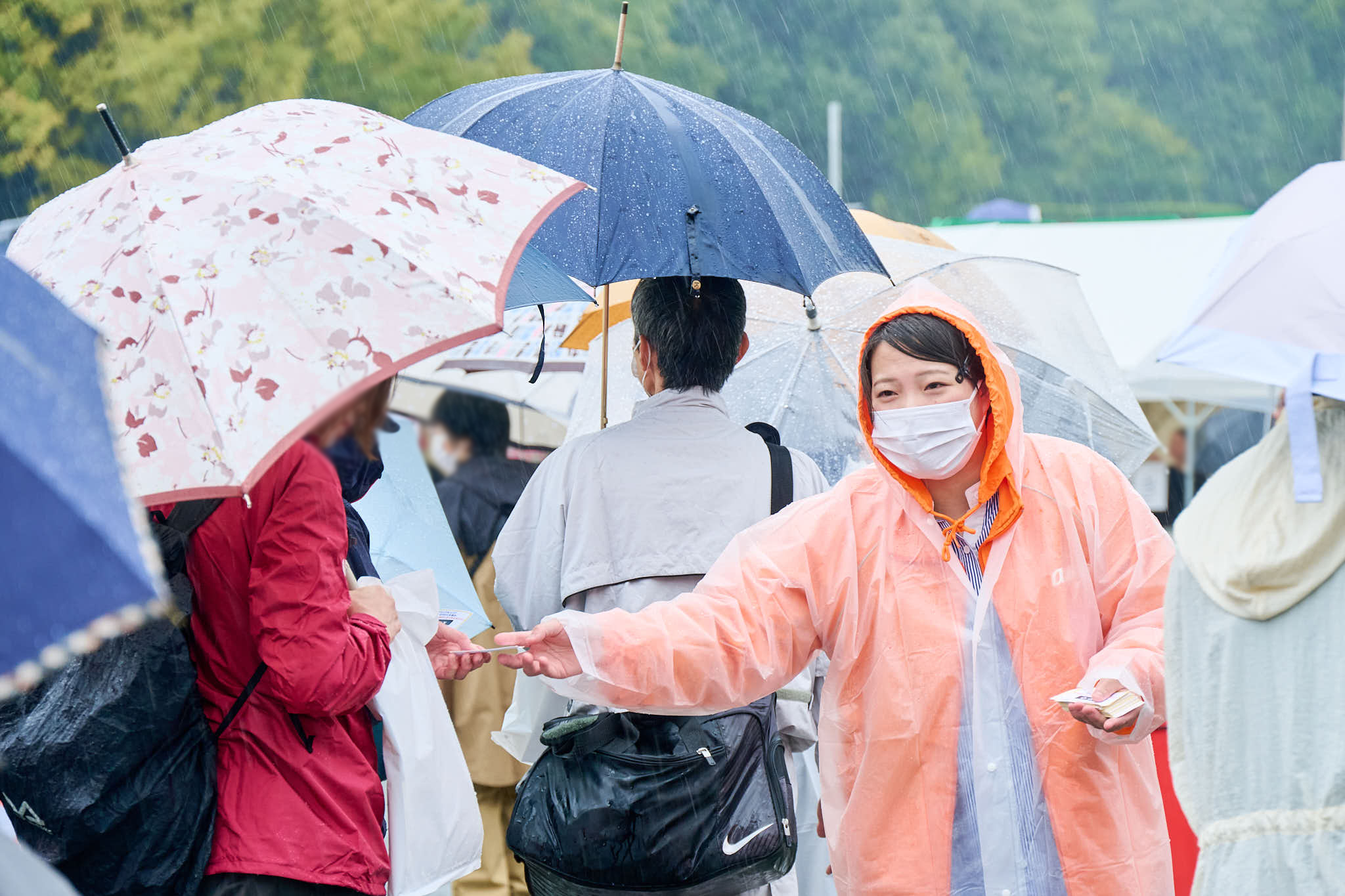 雨の中、傘を差した来場者にスタッフがチラシを配布する様子
