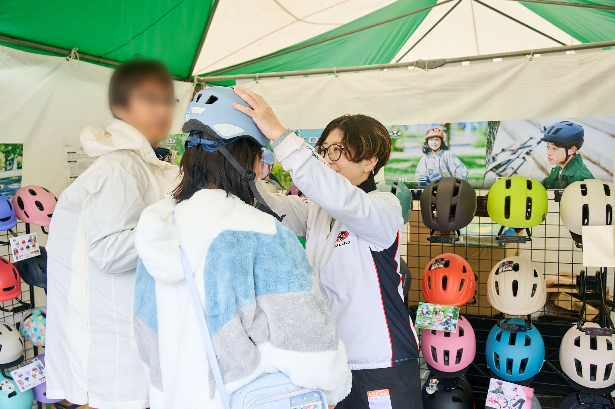 自転車用ヘルメットの展示ブースでスタッフが試着を手伝う様子、カラフルなヘルメットが並ぶ