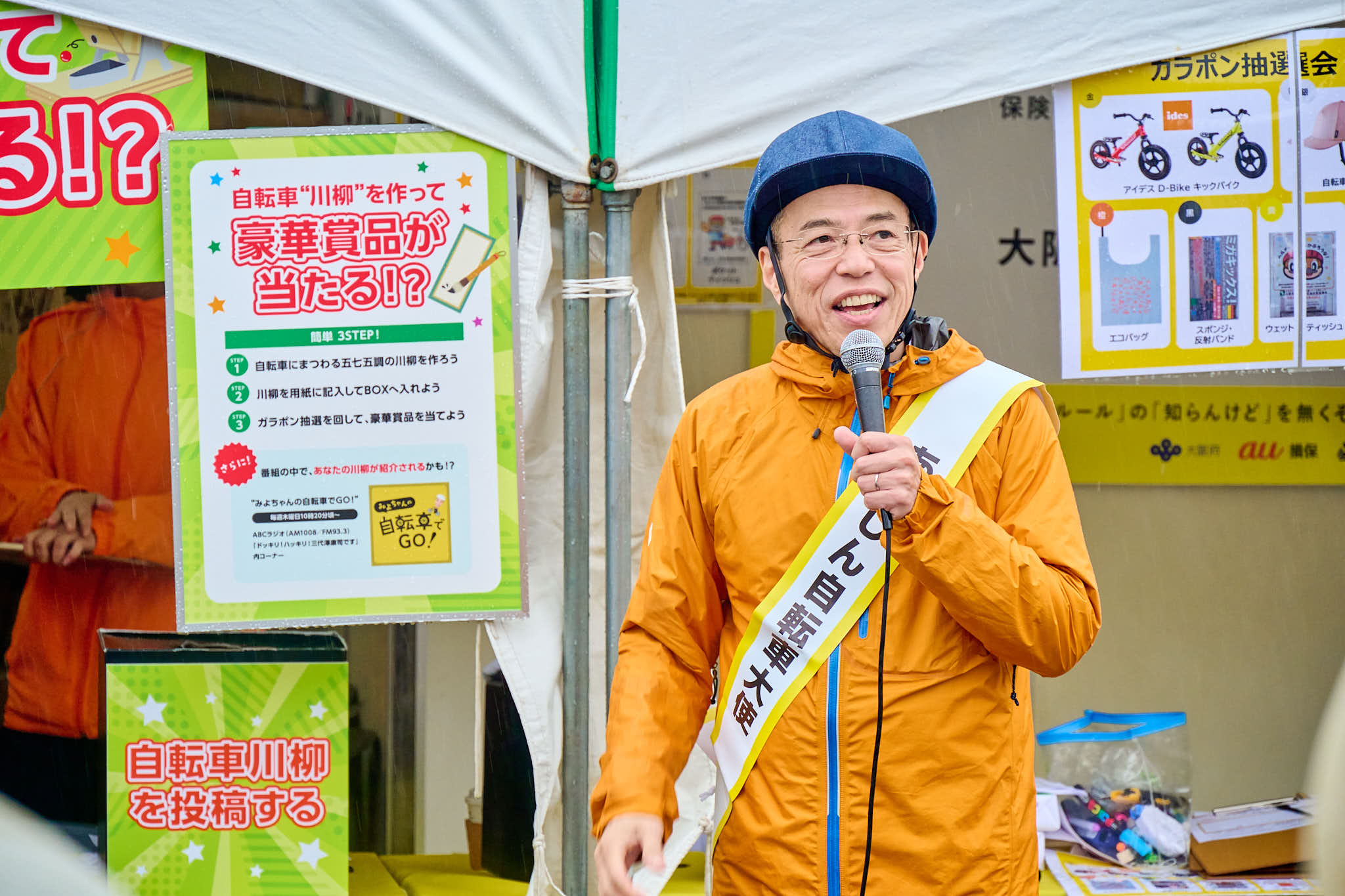 イベントブースで「あんしん自転車大使」みよちゃんがマイクを持ち、参加者とやり取りする様子、背景に景品一覧のポスター