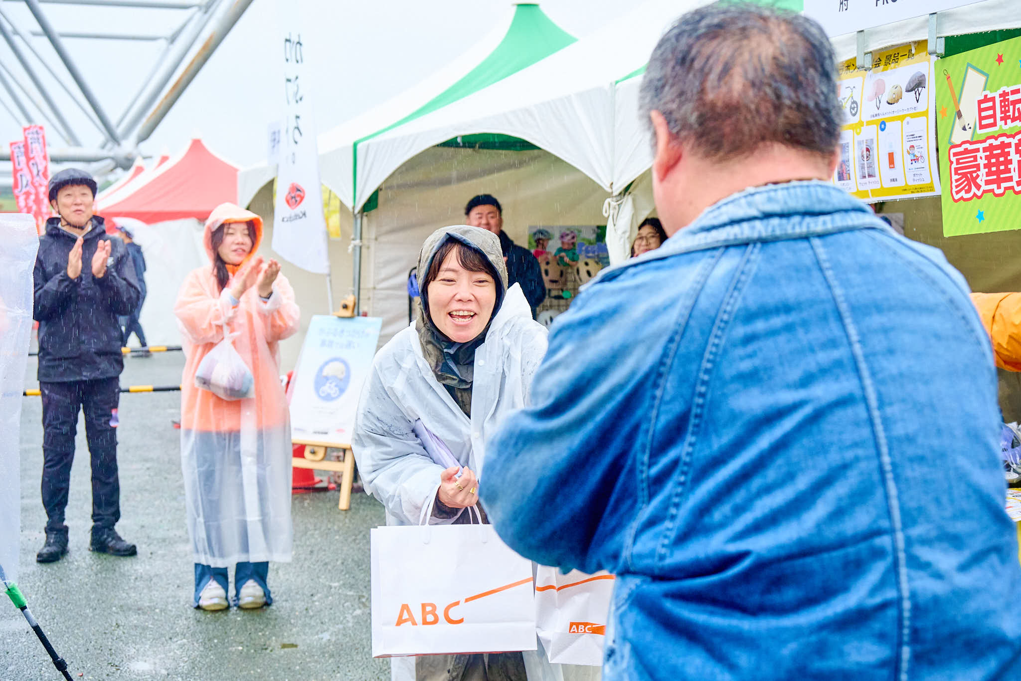 屋外イベントで景品を受け取る参加者とスタッフ、テントの前で拍手する人々