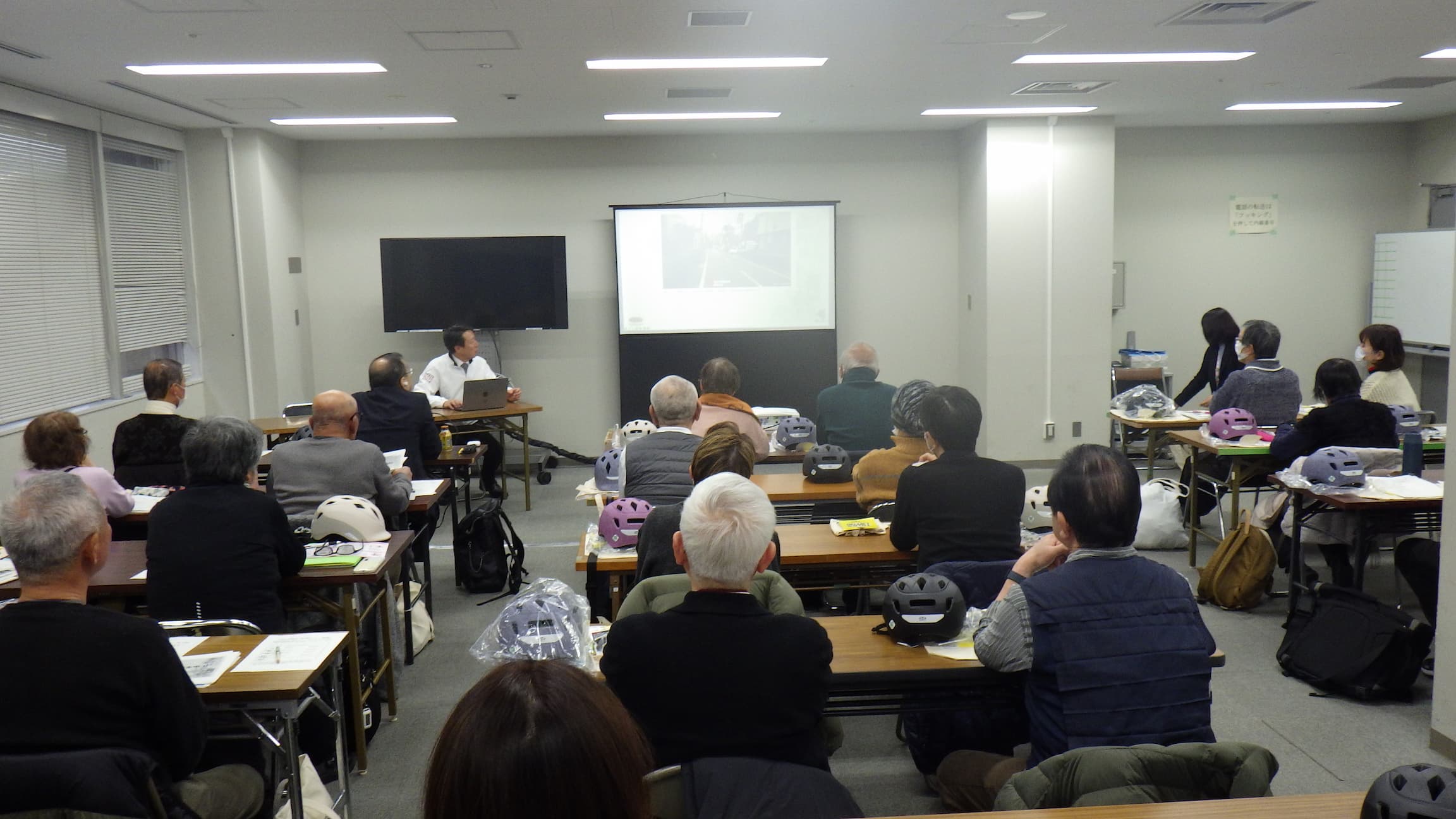 教室で受講者が前方のスクリーンを見ながら講義を受けている様子。机には配布されたヘルメットが置かれている