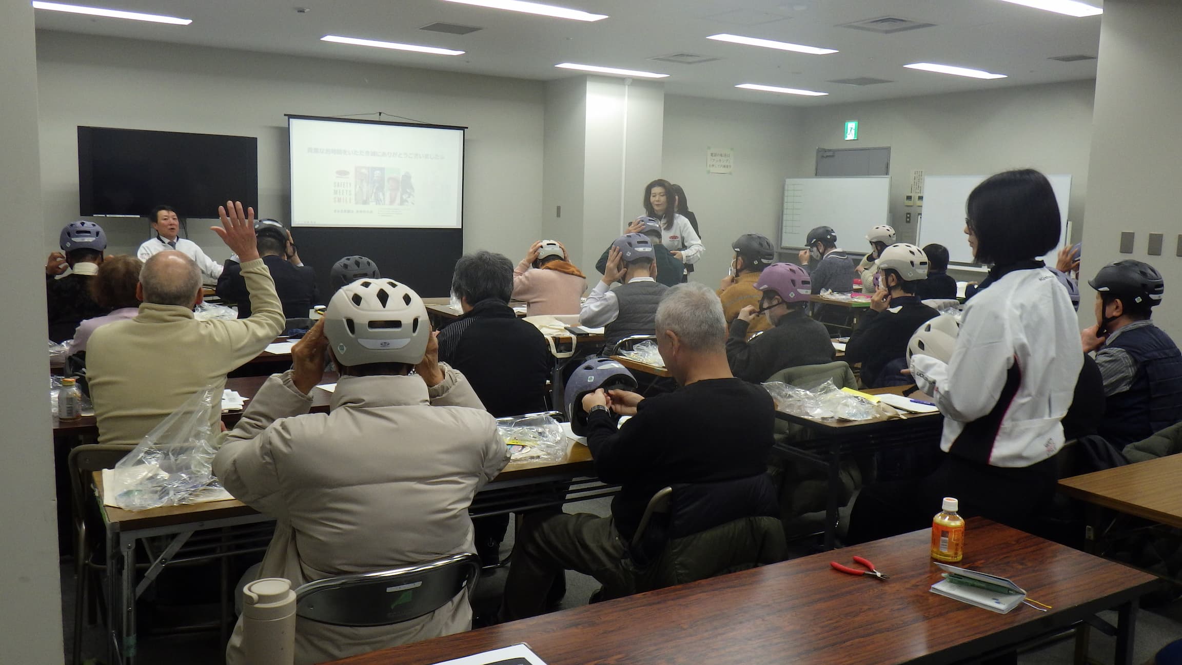 講師が前方で説明する中、受講者が実際にヘルメットを着用して確認している様子