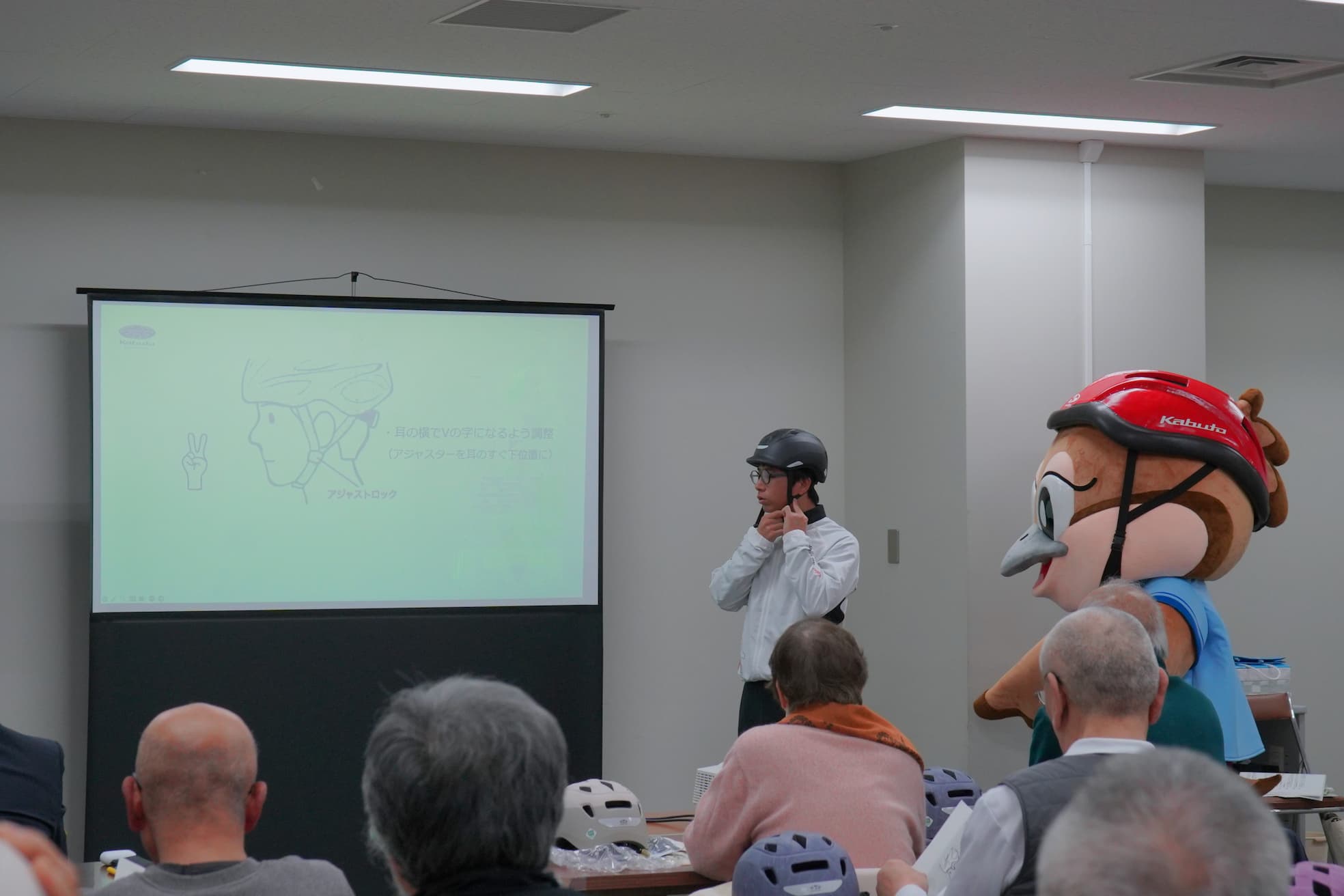 講師がヘルメットの正しいかぶり方を説明し、スクリーンにはヘルメット装着方法のイラストが投影されている。キャラクター(もずやん)も近くで参加している