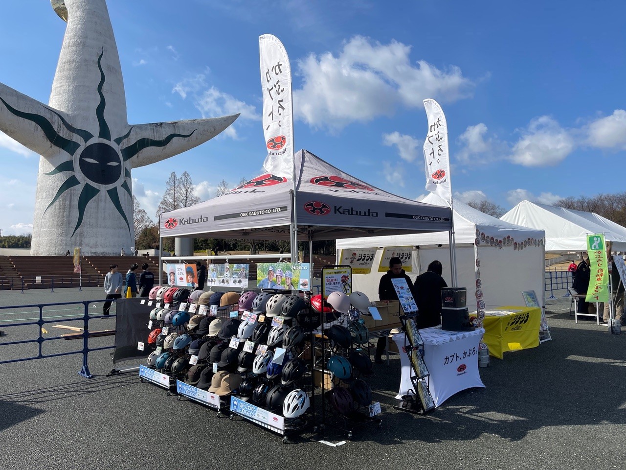 太陽の塔の前に設置された自転車ヘルメット販売ブース。多くのヘルメットが棚に並び、のぼり旗が立っている様子。