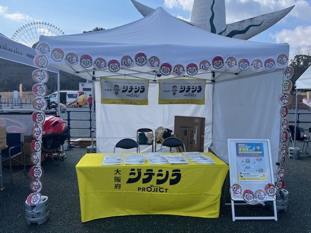 太陽の塔の前に設置された大阪府「ジテシラ PROJECT」の案内テント。黄色いテーブルクロスと掲示物が並んでいる様子。