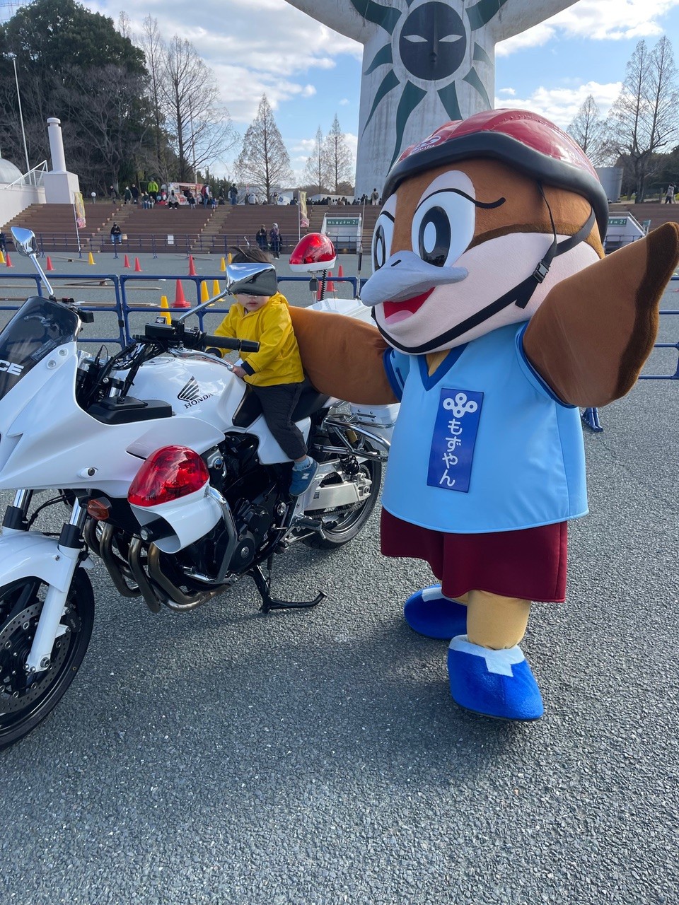白いバイクの横で、ヘルメットをかぶったマスコットキャラクター(もずやん)が腕を広げて立っている様子。バイクには子どもがまたがっている。