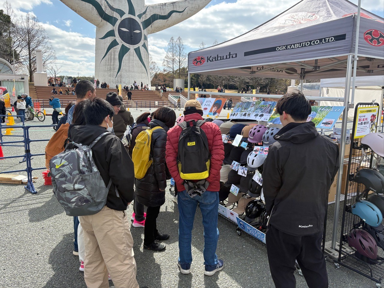 太陽の塔の前で、来場者たちが自転車用ヘルメットの展示ブースを見ている様子。
