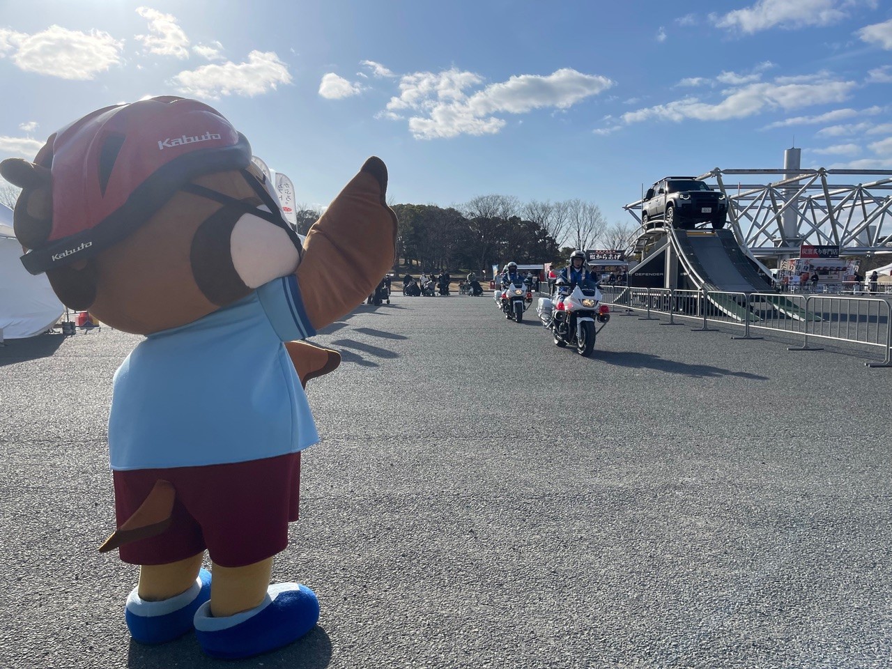 広場で「もずやん」が白バイ走行エリアの方向に手を向けている様子。背景にバイク走行デモ会場が見える。