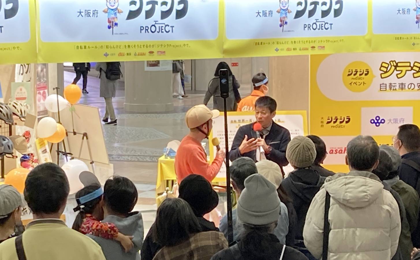 屋内イベント会場で、マイクを持った登壇者2人が黄色のブース前で来場者の前に立ち、周囲に大勢の人が集まっている様子