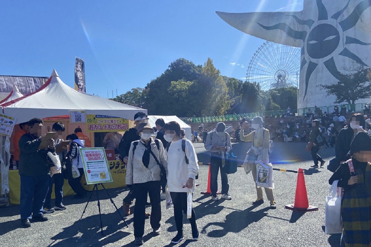 屋外イベント会場でテントブースの前に人が並び、背景には大きな観覧車とステージ装飾が見える様子