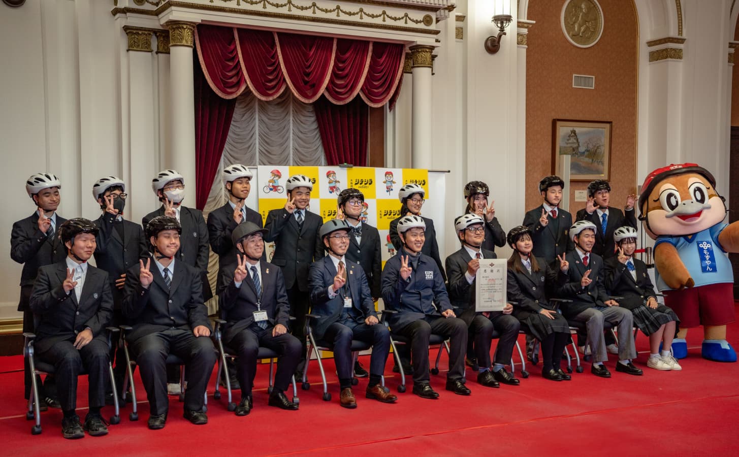 屋内の式典会場で、自転車用ヘルメットを着用した学生と関係者が並び、表彰状を手に記念撮影をしている。右側にマスコットキャラクター(もずやん)が立ち、背後にはチェック柄のパネルと赤いカーテンがある。
