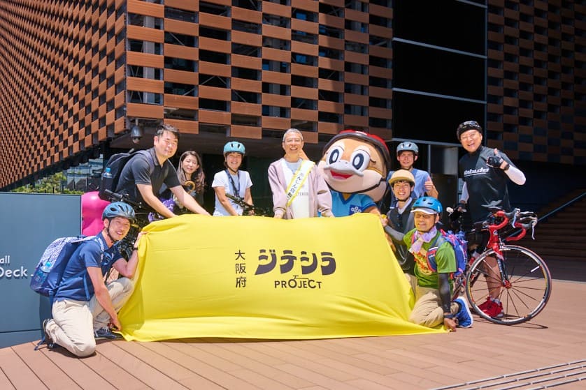 屋外のウッドデッキで、自転車用ヘルメットを着用した複数人が黄色いカバーをかけた台を囲んで並び、記念撮影をしている。カバーには『大阪府 ジテシラ PROJECT』と書かれており、右側に自転車とマスコットキャラクターがある。